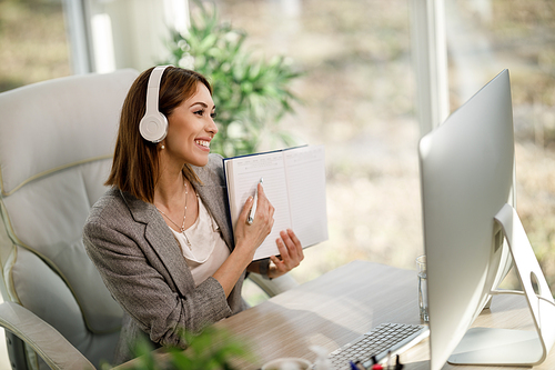 A cheerful young woman sitting alone in her home office with headphones and using a computer to have a web conference.