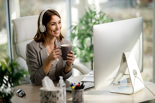 A cheerful young woman with headphones enjoying a cup of coffee while working on computer at the home office.