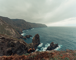 A landscaped shot of the cliffs and giant stones in europe with the wild ocean breaking against it on green and brown tones