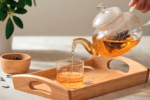A hand pouring tea from glass teapot on wooden serving tray