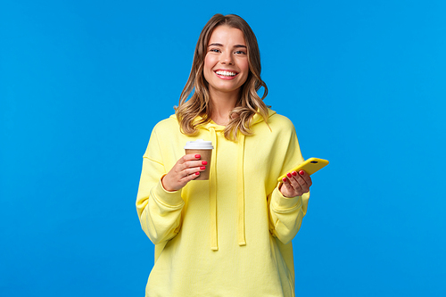 Cheerful young female student standing near cafe ordered take-away coffee, drinking from paper cup and holding mobile phone, look at camera with beaming white smile, satisfied.