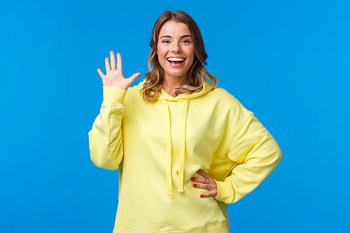 Cheerful friendly outgoing blond girl waving hand, informal greeting concept, smiling white teeth saying hi, welcoming new members to company, see friend, stand blue background.