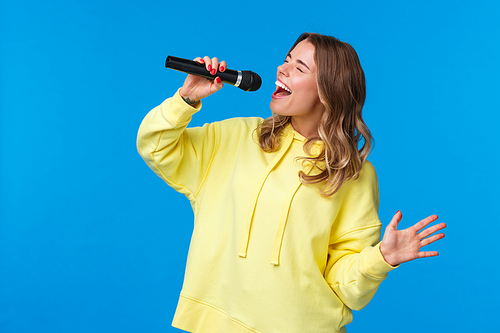 Carefree happy blond european girl having fun, enjoy singing karaoke with all heart and passion, close eyes holding microphone, standing blue background joyful. Copy space