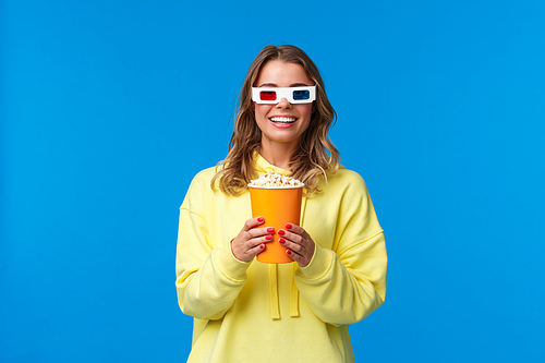 Lets watch movie. Excited cute young blond girl in 3d glasses, holding popcorn and smiling amused as enjoying premiere of film in cinema, standing entertained over blue background.