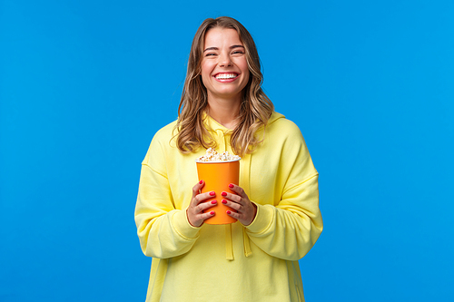 Leisure, fun and youth concept. Friendly happy blond european woman enjoying weekends, eating popcorn and watching comedy, visit cinema to see new movie, stand blue background.