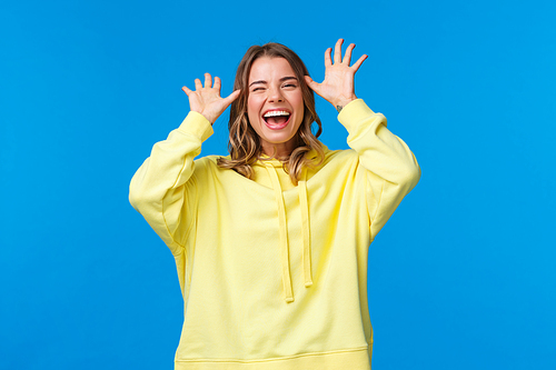 Try catch me. Alluring carefree european female with blond hair having fun, showing funny faces, laughing and smiling joyful, playing, make childish grimaces mocking someone, blue background.