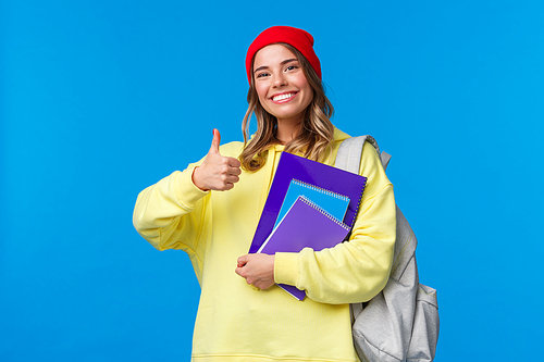 Sounds good. Cheerful cute and smart caucasian girl in red beanie studying hard for exams, encourage student keep up, show thumbs-up in like or approval, hold notebook and backpack.
