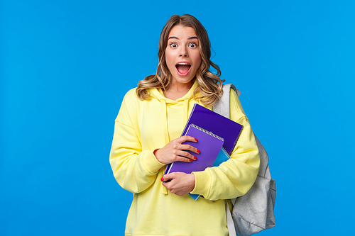 Amused and happy excited pretty blond girl in yellow hoodie, gasping and looking amazed as hear great news, holding notebooks and carry backpack at college, gossiping, blue background.