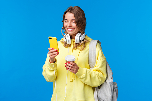 Girl using mobile phone to contact friend after college, texting as walking along street with backpack, take-away coffee cup and headphones, smiling joyfully smartphone display.