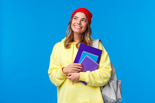 Dreamy cute smart college girl, female student in hipster red beanie, yellow hoodie, looking thoughtful upper left corner imaging summer holidays, smiling holding notebooks and backpack.