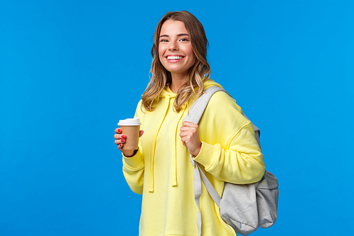 Joyful young blond female student with backpack smiling camera satisfied as holding cup of take-away coffee from favorite cafe after classes, drinking beverage, stand blue background.