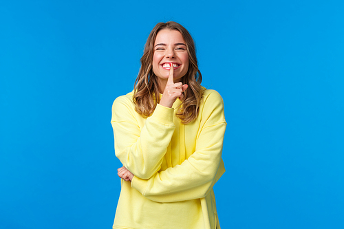 Happy cheerful silly blond caucasian woman in yellow hoodie hiding secret, prepare b-day surprise for boyfriend, shush with index finger pressed to lips as smiling joyfully, blue background.