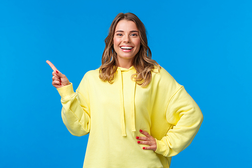 Take a look. Upbeat good-looking blond girl pointing finger left, recommend promo, advertise product, look pleased and smiling camera, explain project, standing blue background.
