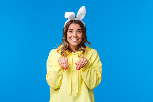 Holidays, traditions and celebration concept. Cute and silly smiling blond woman lovely looking at camera and mimicking rabbit with fake ears on head, standing blue background.