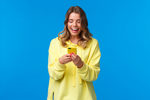 Technology and people concept. Happy smiling blond girl using mobile phone in yellow case, messaging look at smartphone display with pleased expression, send funny meme, blue background.
