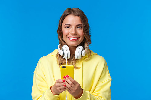 Close-up portrait of young blond european hipster girl with blond short hair and headphones over neck, holding smartphone, texting message, use mobile phone, blue background. Copy space