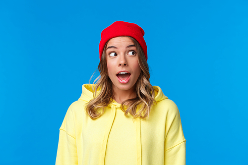 Excited good-looking caucasian female student in red beanie and yellow hoodie, look upper right corner and open mouth fascinated, hear interesting gossip, standing blue background.