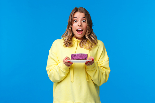Celebration, party and lifestyle concept. Excited and happy cheerful blond girl on diet eating tasty fruit cake, looking surprised and joyful camera, standing blue background.