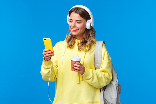 Carefree smiling blond woman in yellow hoodie listening music in headphones, picking song from playlist as drinking coffee on her way home from university, hold backpack, blue background.