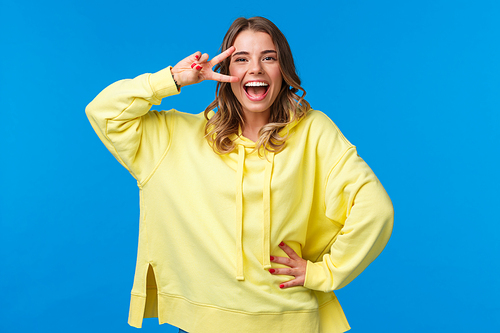 Portrait of happy optimistic laughing girl with blond short hair, smiling joyfully as show disco peace gesture near eye, posing for photo near blue background, having fun.