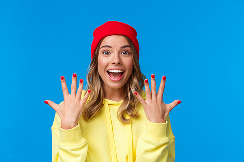 Portrait of amused good-looking blonde hipster girl smiling joyfully, explain something showing number ten, tenth with hands and look camera upbeat, order dozen, stand blue background.