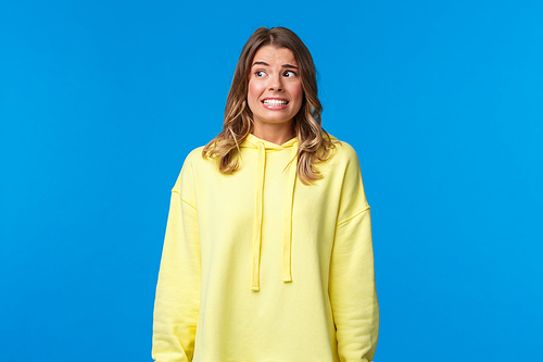 Anxious and embarrassed, alarmed insecure girl have panic and nervous thoughts of someone found out her dirty little secret, smile awkward and worried look around, stand blue background.