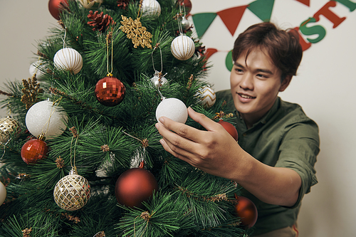 handsome man decorating christmas tree with baubles and looking at camera at hom
