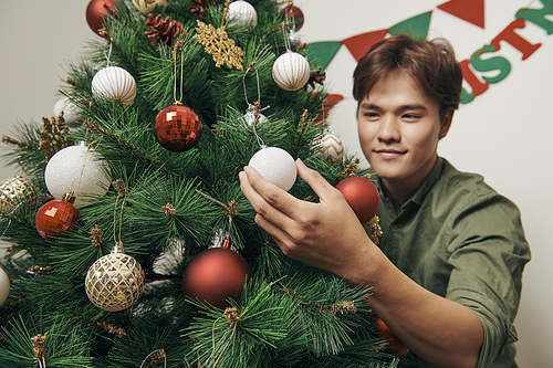 handsome man decorating christmas tree with baubles and looking at camera at hom