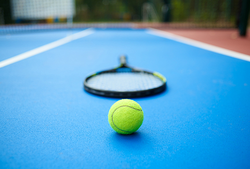 Frontview of yellow tennis ball is laying near professional racket on blue carpet of opened tennis cort. Contrast image with satureted colors and shadows. Concept of sport equipment photo.