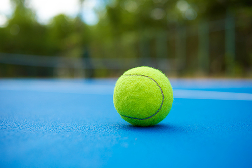 Yellow ball is laying on blue tennis court background. Blurred green plantings and trees behind. Contrast image with satureted colors. Concept of sport equipment photo.