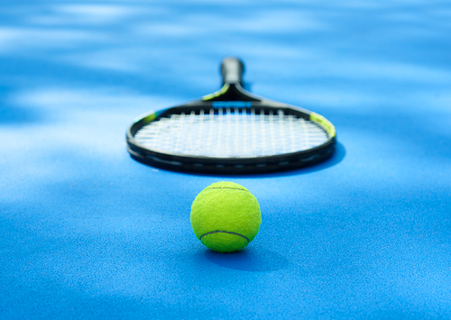 Yellow tennis ball is laying near professional racket on blue cort carpet. Made for playing tennis. Contrast image with satureted colors and shadows. Concept of sport equipment photo.