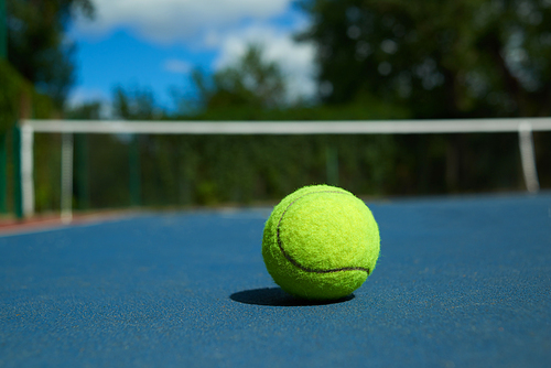 Frontview of yellow bright tennis ball is lying on on blue carpet of opened court during sunny day. Made for playing tennis. Contrast image with satureted colors. Concept of tennis outfit photografing