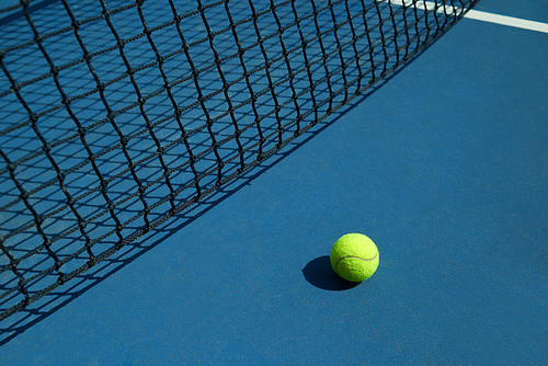 Yellow tennis ball is laying near black opened tennis court's net. Contrast image with satureted colors and shadows. Concept of tennis outfit photografing.