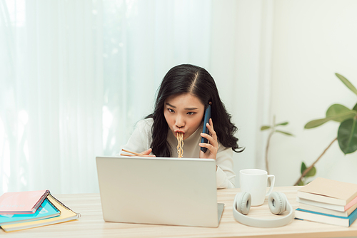 woman eating noodles while working with laptop and cellphone in office