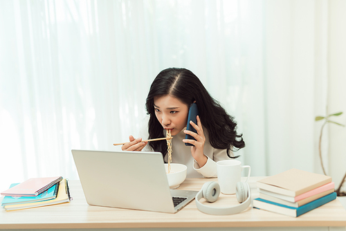 Confident smiling attractive young asian woman entrepreneur having lunch break while sitting at the office desk and using mobile phone