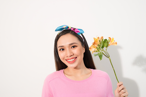 Close up portrait of adorable, beautiful, cute woman in casual clothes with beaming smile and long blond hair, holding three flowers in hand near cheek, standing over white background