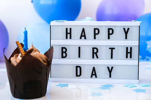 Lightbox with text HAPPY BIRTHDAY and chocolate cupcake candle on Abstract defocused blurred festive background for holiday. Blue ballons and confetti, birthday cake. Greeting card Boy concept