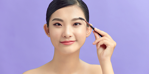 Young woman smiling and using brow brush isolated over purple background