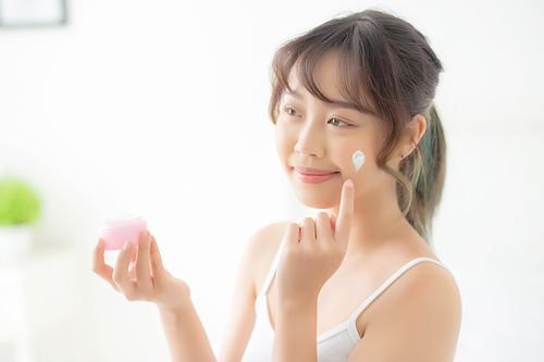 beautiful young asian woman happy applying cream or lotion with moisturizer to skin face, beauty asia girl applying skincare touch facial with cosmetic makeup, healthy and wellness concept.