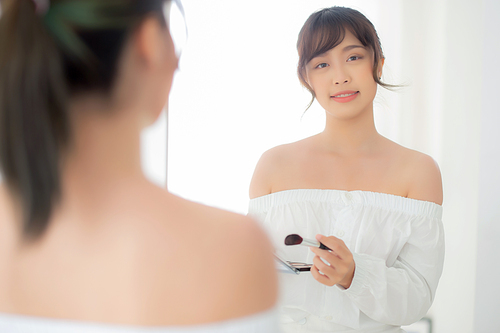 Beauty portrait young asian woman smiling with face looking mirror applying makeup with brush cheek in the bedroom, beautiful girl holding blusher, skin care and cosmetic table fashion concept.