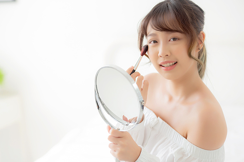 Beauty portrait young asian woman smile with face looking mirror applying makeup with brush cheek in the bedroom, beautiful of girl holding blusher, skin care and cosmetic concept.