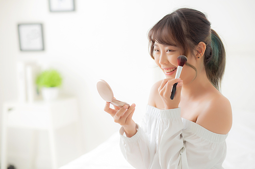 Beauty portrait young asian woman smiling with face looking mirror applying makeup with brush cheek in the bedroom, beautiful girl holding blusher facial, skin care and cosmetic concept.