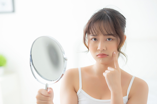 Beautiful young asian woman looking the mirror with acne problem at bedroom, trouble of beauty on face, zit treatment, asia girl is pimple having worry and displeased, skincare and healthy concept.