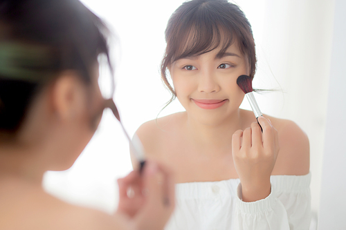 Beauty portrait young asian woman smile with face looking mirror applying makeup with brush cheek in the bedroom, beautiful of girl holding blusher, skin care and cosmetic concept.