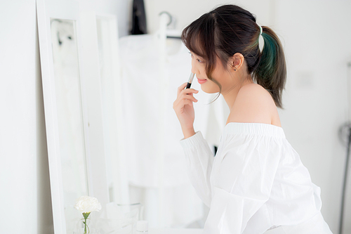 Beauty portrait young asian woman smile with face looking mirror applying makeup with brush cheek in the bedroom, beautiful of girl holding blusher, skin care and cosmetic concept.