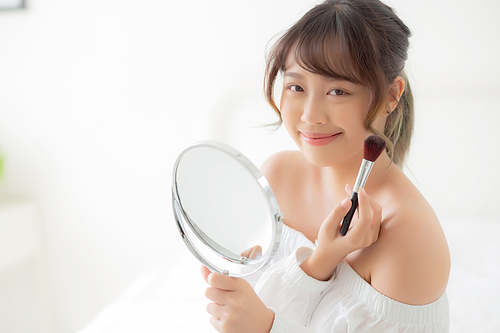 Beauty portrait young asian woman smile with face looking mirror applying makeup with brush cheek in the bedroom, beautiful of girl holding blusher, skin care and cosmetic concept.