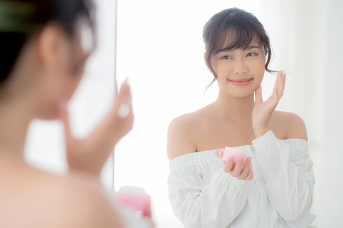 Beautiful portrait young asian woman applying cream moisturizer or lotion skin care cosmetic on face looking mirror, girl with treatment facial, health and wellness concept.