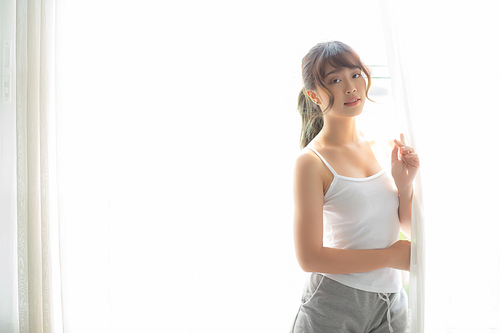 Portrait of beautiful young asian woman standing the window and smile while wake up with sunrise at morning, girl happy with freshness and cheerful, lifestyle and relax concept.