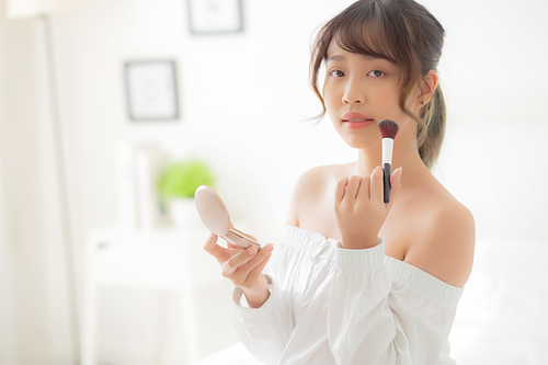 Beauty portrait young asian woman smiling with face looking mirror applying makeup with brush cheek in the bedroom, beautiful girl holding blusher facial, skin care and cosmetic concept.