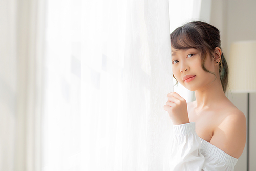 Portrait of beautiful young asian woman standing the window and smile while wake up with sunrise at morning, girl happy with freshness and cheerful, lifestyle and relax concept.